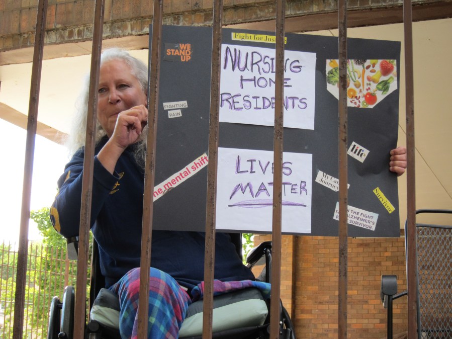 A white woman with long white hair in a wheelchair holds up a protest sign that says "Nursing Home Residents Lives Matter" - she is behind a metal fence, which encloses her in her facility