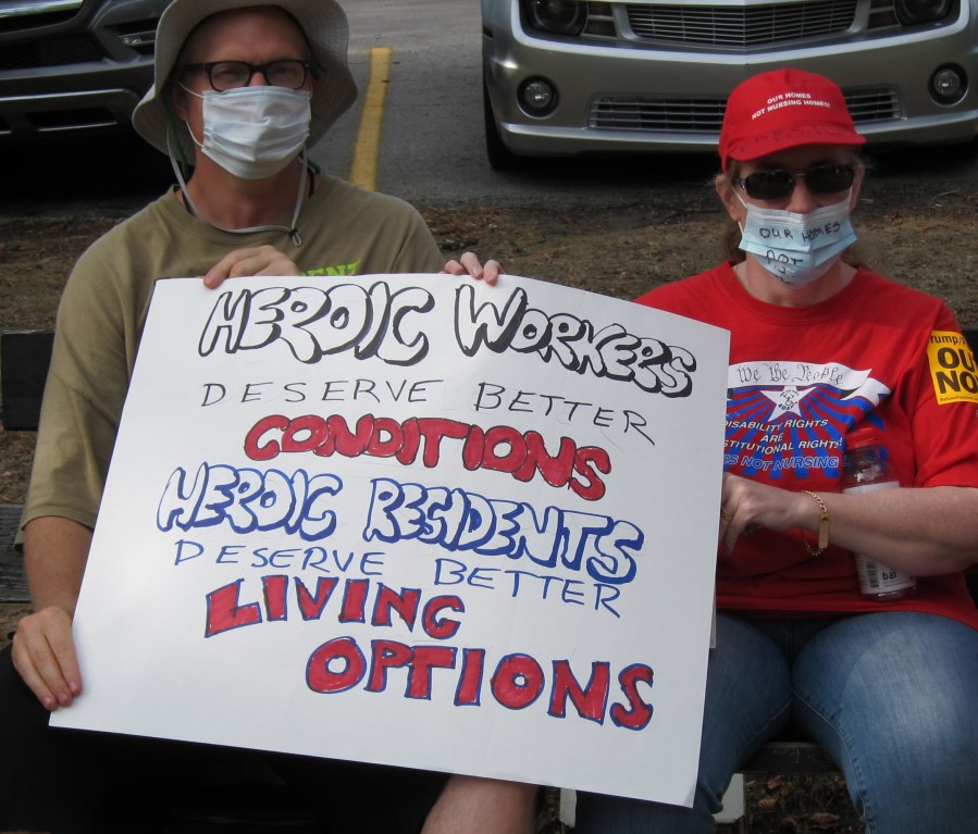 Two white people in surgical masks and glasses hold a sign that says "Heroic workers deserve better conditions, heroic residents deserve better living options"